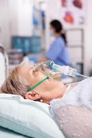 Senior woman contaminated with covid19 wearing oxygen mask in clinic. Patient in hospital room laying on medical bed with infected lung during coronavirus pandemic having respiratory problem. photo