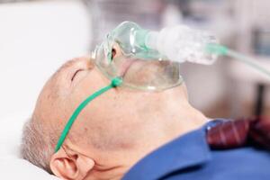 Senior man doing inhalation through oxygen mask because of covid19. Patient in hospital room laying on medical bed with infected lung during coronavirus pandemic having respiratory problem. photo