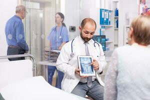 Healthcare specialist explaining senior woman diagnosis of braing trauma on tablet pc. Medical therapist showing and talking about x-ray with patient during consultation. photo