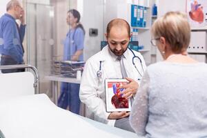 Doctor talking about efect of heart attack pointing at tablet pc during senior woman examination. Cardiologist wearing stethoscope discussing with patient about unhealthy heart disease. photo