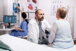 Healthcare specialist using stethoscope in private clinic using stethoscope. Medical practitioner examining lungs of caucasian in modern clinic dressed in white coat. photo