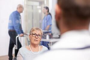 Disabled old person in wheelchair during treatment with geriatrician doctor. Medical specialist wearing stethoscope discussing with invalid elderly woman in consultation room of private. photo