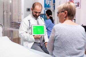 Mature woman in hospital consultation room listening doctor with tablet green screen. Ready chroma mockup for your app, text, video or other digital asset. photo