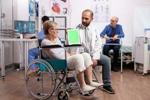 Doctor using tablet pc with green screen while consulting handicapped senior woman in wheelchair. Medical practitioner using device with chroma key during coonsultation of invalid senior woman. photo