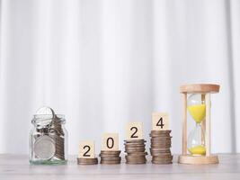 Hourglass and Wooden block with number 2024 on stack of coins. The concept of saving money, Financial, Investment and Business growing in new year 2024. photo