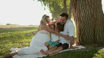 A large family with a pregnant mother hugging while sitting by a tree on the lawn. video