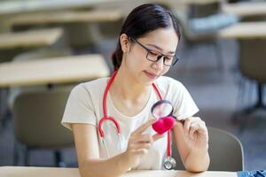 Medical student holding a magnifying glass and looks at a yarn red heart on blurred background. Asian medical student with Heart disease concept. photo