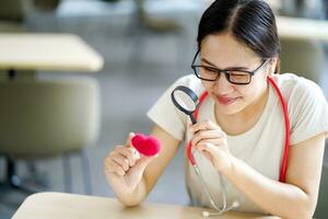 Medical student holding a magnifying glass and looks at a yarn red heart on blurred background. Asian medical student with Heart disease concept. photo