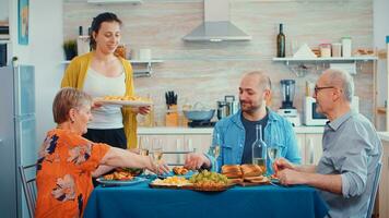 Young wife putting potatoes on the table and clinking a glass of wine with her husband. Caucasian family enjoying time at home, in the kitchen sitting by the table, eating dinner together and drinking photo