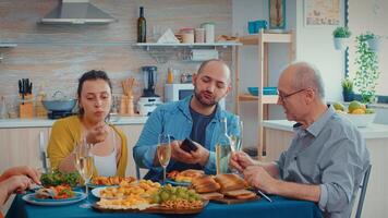 Elderly father watching photos on smartphone of young man while sitting in kitchen during family dinner. Multi generation, two couples talking and eating during a gourmet male, enjoying time at home