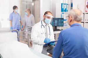 Doctor wearing visor as safety precaution in the course of consultation of senior man. Physician practitioner in the course of private modern clinic appointment during coronavirus pandemic. photo