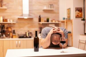 Deprimido hombre teniendo dolor de cabeza después alcohol abuso en cocina. infeliz persona enfermedad y ansiedad sensación agotado con teniendo alcoholismo problemas. foto