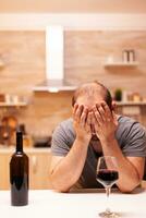 Frustrated drunk young man at home during life crisis with a bottle of red wine. Unhappy person disease and anxiety feeling exhausted with having alcoholism problems. photo
