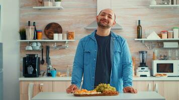 marido chateando en el cocina mientras su familia prepara el cena en antecedentes. retrato contento sonriente joven hombre mirando a cámara, cabeza Disparo retrato, comiendo queso, extendido familia alrededor él foto