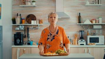 mayor mujer teniendo un vídeo llamada en el cocina mientras su familia prepara el cena en el antecedentes. pov en línea Internet moderno conferencia, charlando, comunicación, charla conversacion llamada vía cámara web foto