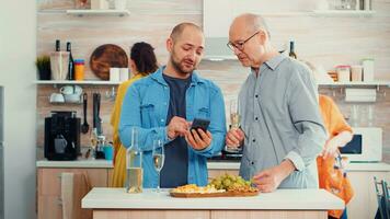 Young man showing photos on smartphone to his father. Group of mixed generations people sitting around table in a modern kitchen and using mobile phone while women prepare the meal.