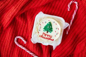 A festive Christmas cupcake with a white frosting and a green tree on top, on a red knitted background with candy canes. Merry Christmas lettering. photo