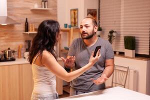 marido atrapado engañando teniendo un conflicto con esposa en hogar cocina. calentado enojado frustrado ofendido irritado acusando su hombre de infidelidad demostración él mensajes en teléfono inteligente Gritando desesperado. foto