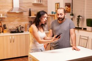 Wife screaming at unfaithful husband in the kitchen. Jealous woman cheated angry frustrated offended irritated accusing her man of infidelity showing him messages on smartphone yelling desperate. photo