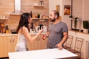 Woman looking disappointed at husband after catching him cheating while holding phone. Heated angry frustrated offended irritated accusing her man of infidelity showing him messages . photo