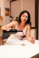 Unhapy woman pouring red wine from bottle in glass sitting alone at table in kitchen. Unhappy person disease and anxiety feeling exhausted with having alcoholism problems. photo