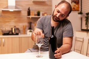 Alcoholic man being depressed and frustrated holding bottle of red wine in kitchen. Unhappy person disease and anxiety feeling exhausted with having alcoholism problems. photo
