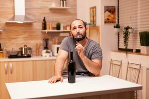 Depressed young man with alcohol addiction having wine degustation in kitchen sitting at table. Unhappy person disease and anxiety feeling exhausted with having alcoholism problems. photo