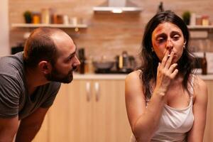 Woman suffering after husband beating and smoking sitting on chair in kitchen. Violent aggressive man abusing injuring terrified helpless, vulnerable, afraid, and panicked woman. photo