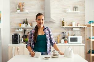 Woman holding cup of tea during morning in kitchen while enjoying breakfast. Preparing tea in the morning, in a modern kitchen sitting near the table. Putting with hands, healthy herbal in pot. photo