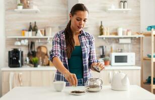 Housewife using aromatic herbs to make hot natural tea for enjoying breakfast. Preparing tea in the morning, in a modern kitchen sitting near the table. Putting with hands, healthy herbal in pot. photo