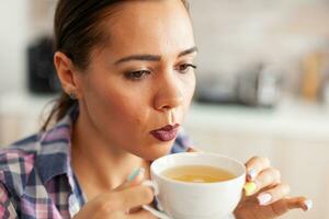 Close up of woman in kitchen trying to drink hot green tea with aromatic herbs. Pretty lady sitting in the kitchen in the morning during breakfast time relaxing with tasty natural herbal tea. photo