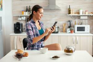 Cheerful woman using smarthphone in kitchen during breakfast and armoatic green tea. Holding phone device with touchscreen using internet technology scrolling, searching on intelligent gadget. photo