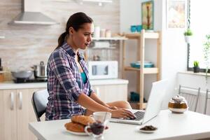 mujer trabajando desde hogar utilizando ordenador portátil en cocina durante desayuno. trabajando desde hogar utilizando dispositivo con Internet tecnología, hojeada, buscando en artilugio en el Mañana. foto