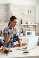 Woman looking at laptop in kitchen holding cup with green tea in the morning. Working from home using device with internet technology, browsing, searching on gadget in the morning. photo