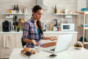 Stresed woman using laptop in kitchen during breakfast with cup of green tea. Working from home using device with internet technology, browsing, searching on gadget in the morning. photo