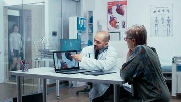 Young male doctor explaining possible heart disease to elderly retired old woman patient. Heart disease problems presented by cardiologist cardiology, heart attach. Health care in modern private clinic. Medical staff diagnostic photo