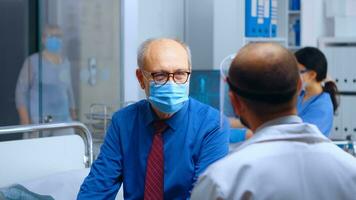 Physician talking with patient in mask during COVID 19 health crisis. Medical healthcare system global pandemic. Medicine worker offering consultation to elderly retired old man. Handheld slow motion photo