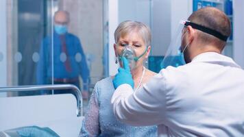 Doctor in mask and visor helping old lady to breathe with a respiratory oxygen mask while sitting on hospital bed in modern private clinic. Coronavirus covid-19 outbreak healthcare crisis. photo
