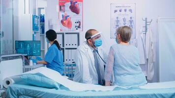 Retired old woman getting a heart check by cardiologist in protective visor and wearing a mask. Healthcare medical medicine consultation, using stethoscope giving professional treatment to sick ill diagnosis from specialist. photo