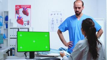 Assistant giving doctor x-ray image while working on computer with green screen monitor in hospital cabinet. Desktop with replaceable screen in medical clinic while doctor is checking patient radiography for diagnose. photo