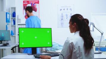 médico tomando notas en computadora con croma llave verde pantalla y enfermero en azul uniforme en hospital gabinete. médico en blanco Saco trabajando en monitor con blanco espacio en clínica gabinete a cheque paciente diagnosticar. foto