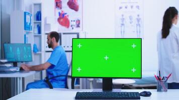 Dolly shot of computer with green screen in hospital cabinet with doctor and assistant working in the background. photo