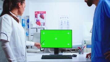 Doctor explaining on computer with green screen in hospital cabinet to nurse in blue uniform. Medic wearing white coat in clinic pointing at desktop with chroma key talking with nurse about patient diagnose photo