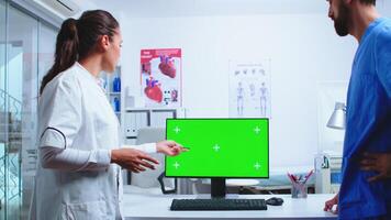 Doctor analysing patient results with assistant on computer with green screen in hospital. Medic wearing white coat in clinic pointing at desktop with chroma key talking with nurse about patient diagnose photo