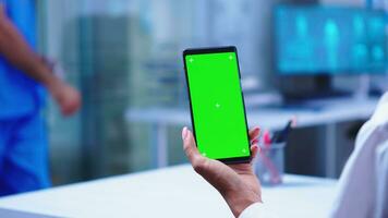 Medic in hospital cabinet holding phone with green screen wearing white coat while nurse opens glass door. Healthcare specialist in hospital cabinet using smartphone with mockup. photo