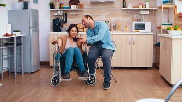 Cheerful disabled woman in wheelchair taking a selfie with husband in kitchen. Hopeful husband with handicapped disabled disability invalid paralysis handicap person next to him, helping her to reintegrate photo