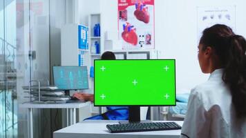 Doctor using computer with green screen mockup in hospital and assistant walks out of cabinet wearing blue uniform. Medic in white coat working on monitor with chroma key in clinic cabinet to check patient diagnose. photo