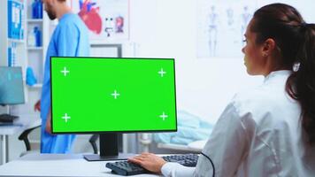 Physician writing diagnose on computer with green screen and assistant wearing blue uniform in the background. Medic in white coat working on monitor with chroma key in clinic cabinet to check patient diagnose. photo