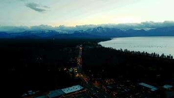 View from a height of the evening city near the sea on the horizon of snowcapped mountains and fluffy clouds. Lanterns burn brightly, the road is illuminated by car lights. Slow traffic on the roads video