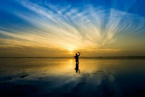 mínimo silueta de contento turista con polos en mano encima cabeza en el playa con amanecer cielo. foto